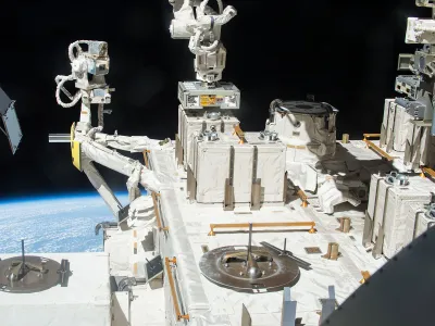 A robotic arm places a container with three panels of bacteria outside the International Space Station.