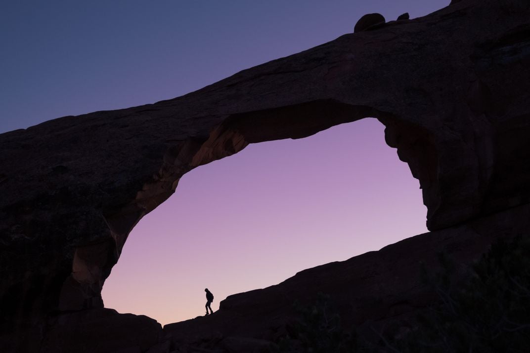 a hiker is seen in silhouette hiking under a rock archway